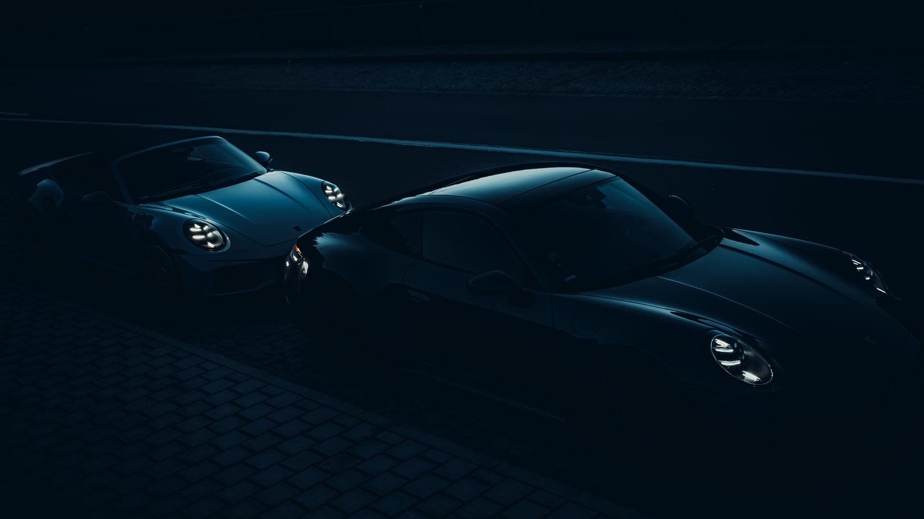Two Porsche 911s facing each other at night on a cobblestone street.